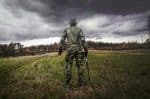 A soldier in camouflage uniform with a rifle stands in a grassy field under a cloudy sky.