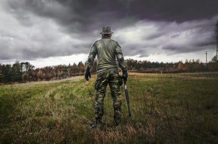 A soldier in camouflage uniform with a rifle stands in a grassy field under a cloudy sky.