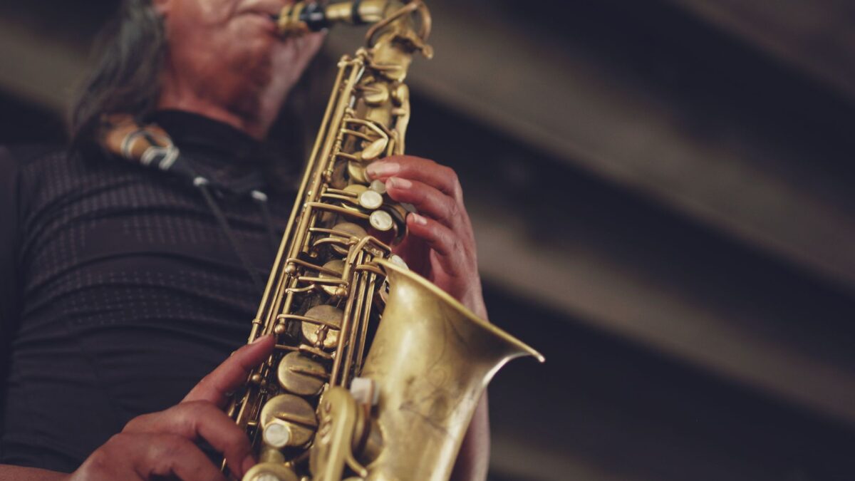 Un muzician cântă la un saxofon alto auriu, fotografiat de jos în sus. Se văd degetele pe clape și o parte din față lângă ambușură, pe un fundal întunecat și estompat.
