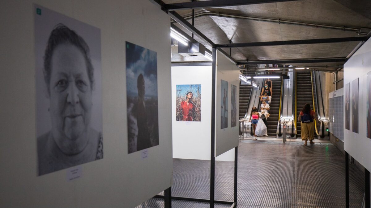 Coridor subteran din beton cu fotografii înrămate, alb-negru și color, expuse pe panouri, un portret alb-negru al unei femei în vârstă în prim-plan și scări rulante în fundal, pe care coboară oameni, unii purtând măști.