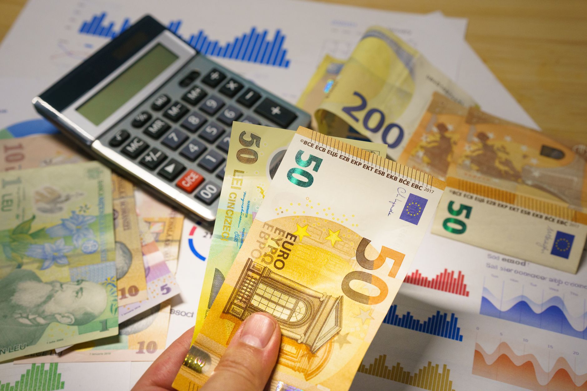 Bancnote de 50 de euro și 50 de lei românești alături de un calculator și grafice imprimate pe hârtie.