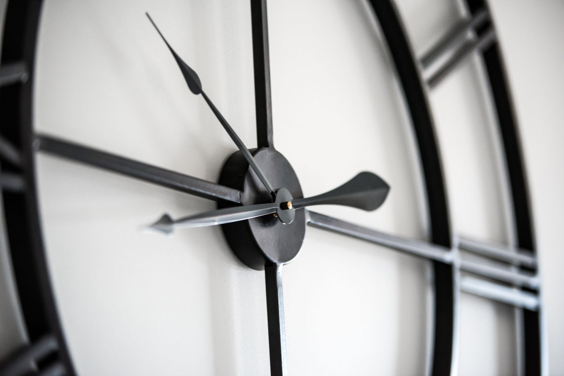 Gros plan sur les aiguilles et le moyeu d’une grande horloge murale métallique noire fixée sur un mur clair.